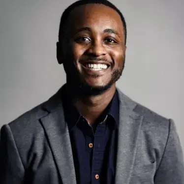 A person with short hair and a beard is smiling at the camera. They are wearing a gray blazer over a dark button-up shirt. The background is plain and softly lit.