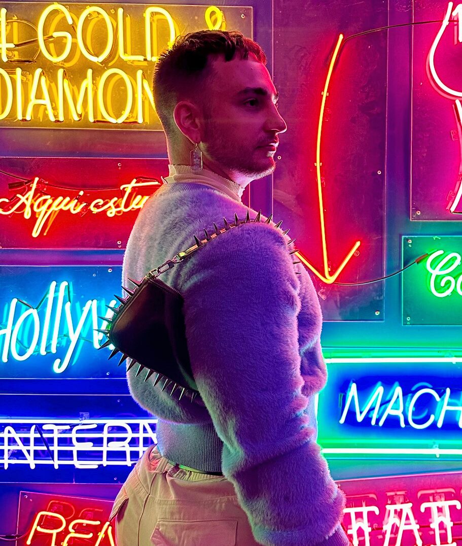 A person wearing a fuzzy lavender sweater with spiked shoulder pads stands in front of a wall covered in colorful neon signs with words like “GOLD & DIAMOND” and “Hollywood.” The neon lights cast bright hues all around.