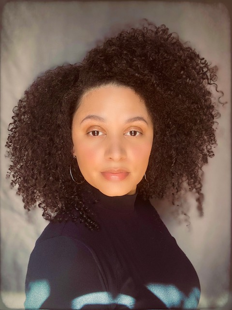 A woman with curly dark hair and hoop earrings poses against a neutral background, wearing a black top and looking directly at the camera with a soft expression.