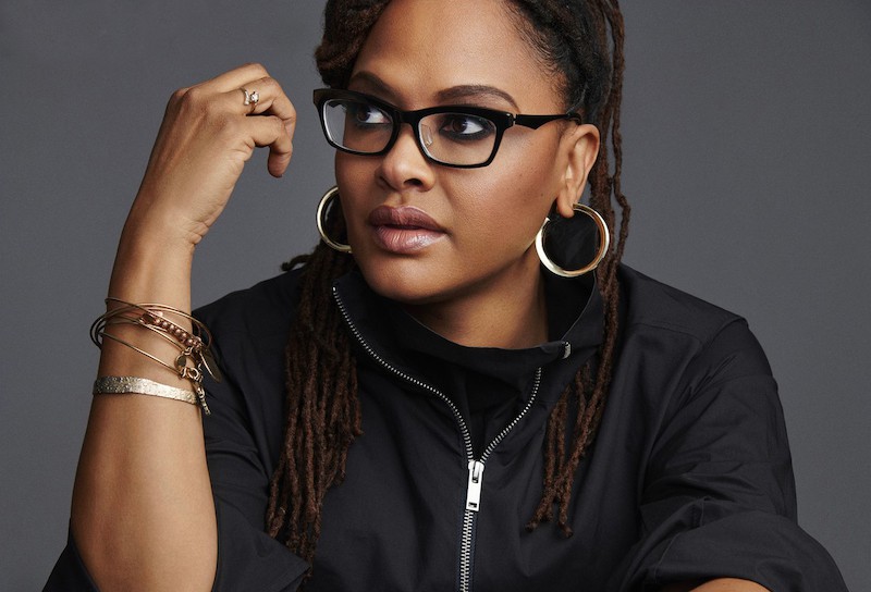 A woman with long dreadlocks resembling Ava DuVernay, wearing black glasses, large gold hoop earrings, and multiple bracelets, poses thoughtfully against a gray background in a black zip-up jacket.
