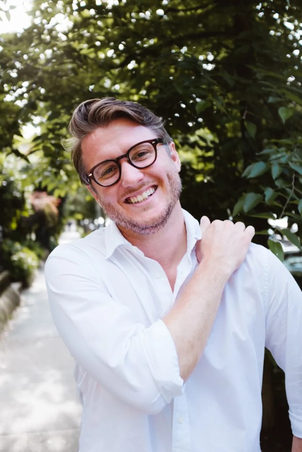 A smiling man with glasses and short, wavy hair stands outdoors on a leafy sidewalk, wearing a white button-down shirt with the sleeves rolled up and one hand resting on his shoulder.