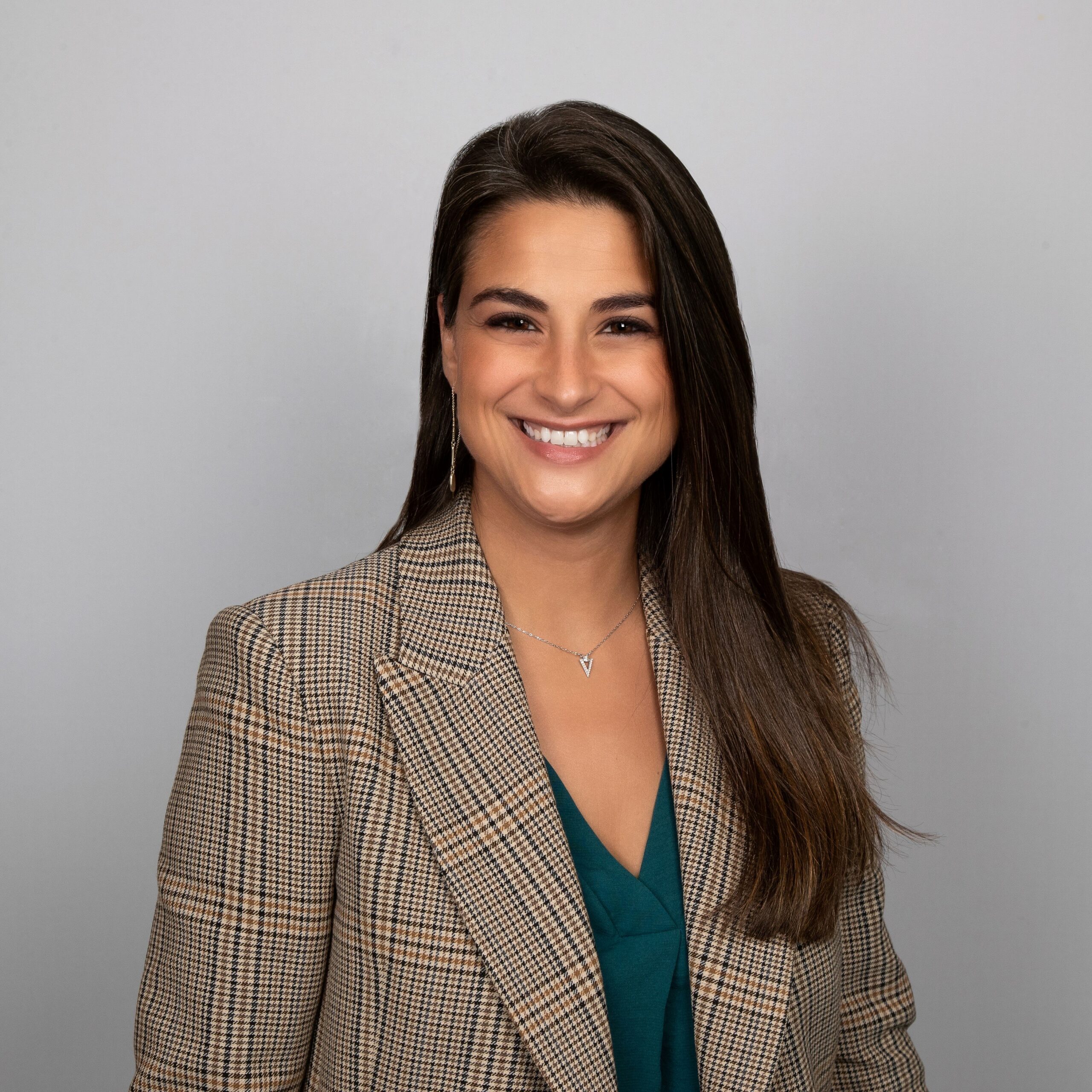 A woman with long brown hair smiles at the camera. She is wearing a brown plaid blazer over a teal top, and a silver necklace. The background is plain light gray.