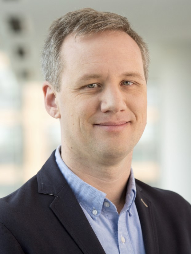 A man with short light brown hair wearing a blue shirt and dark blazer smiles slightly while posing in a bright, modern indoor setting.