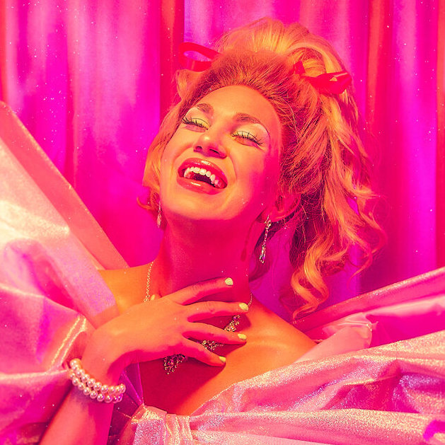 A person dressed in glamorous drag with voluminous blonde hair, sparkling jewelry, and dramatic makeup laughs joyfully in front of bright pink satin curtains, illuminated by vivid pink lighting.