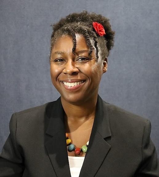 A smiling woman wearing a black blazer, a multicolored beaded necklace, and a red flower in her curly hair, posing in front of a plain gray background.