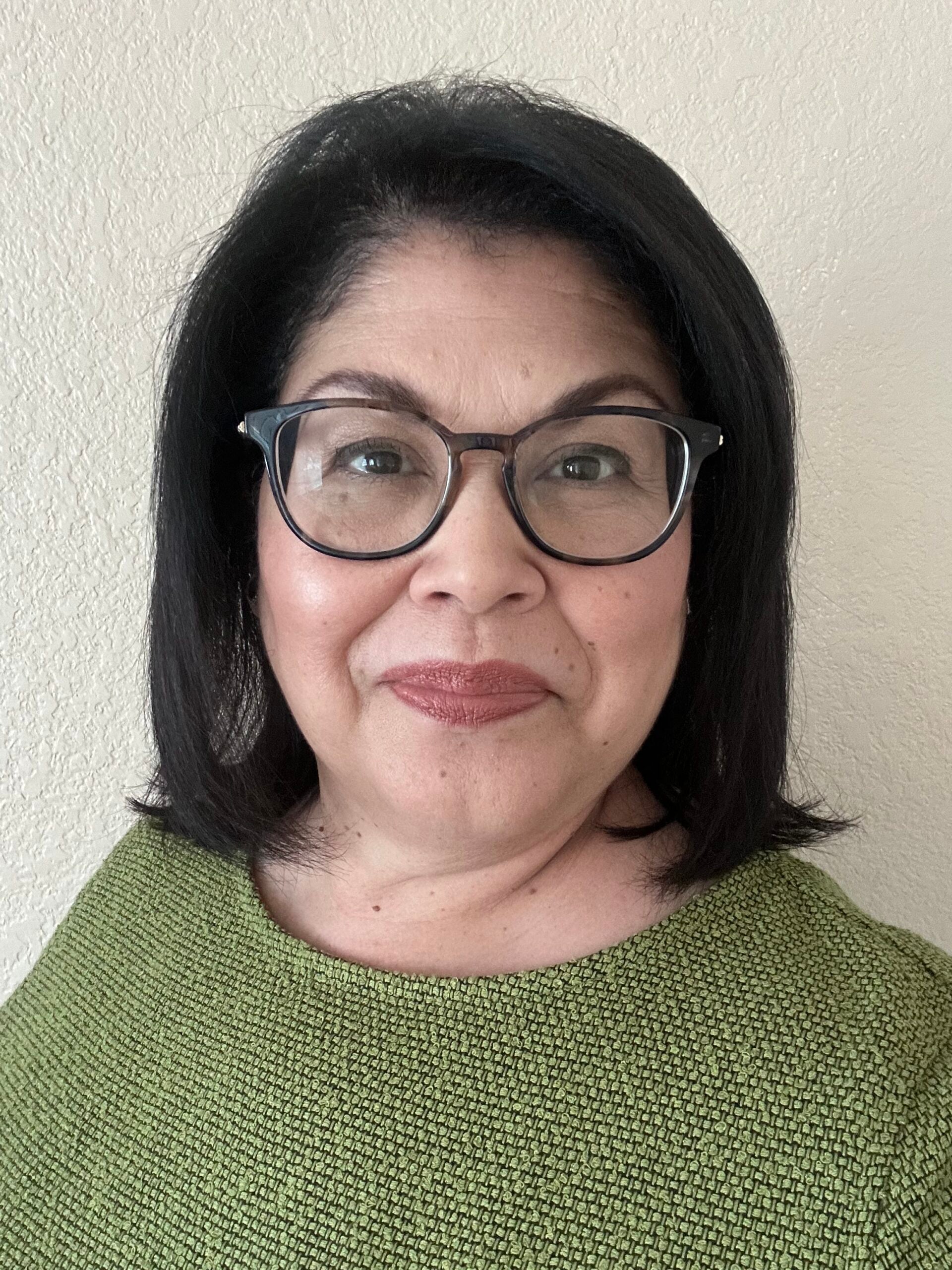A woman with shoulder-length dark hair, wearing glasses and a textured green top, smiles softly while standing in front of a light-colored wall.