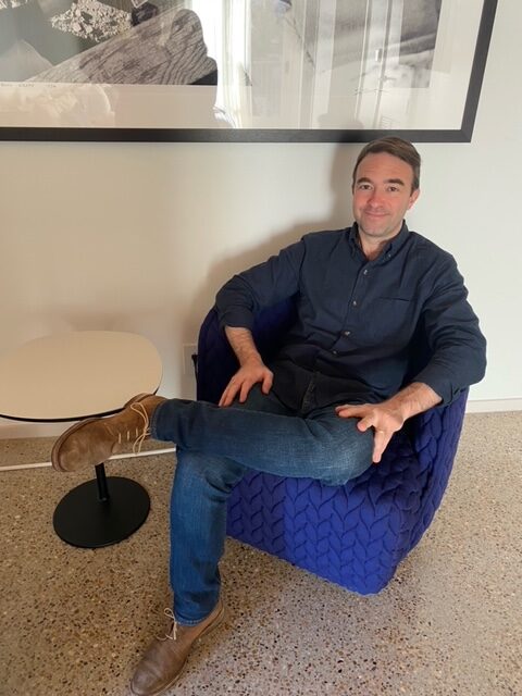 A man in a dark shirt and jeans sits in a blue textured chair with his legs crossed, smiling. He is indoors next to a small round white table and in front of a wall with a black-and-white framed photo.