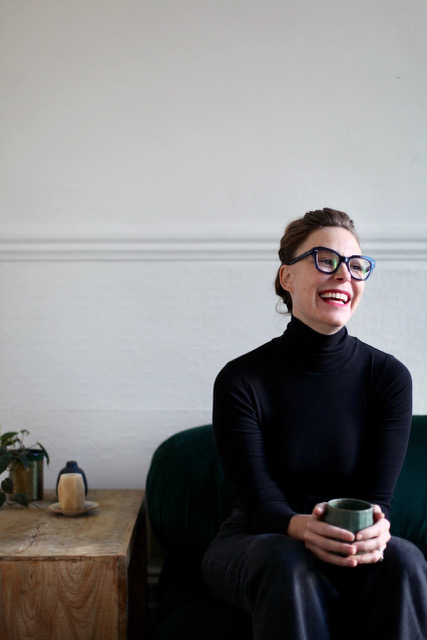 A woman wearing glasses and a black turtleneck sits on a dark green sofa, smiling and holding a mug. A small wooden table with a plant and teapot is beside her, against a white wall background.
