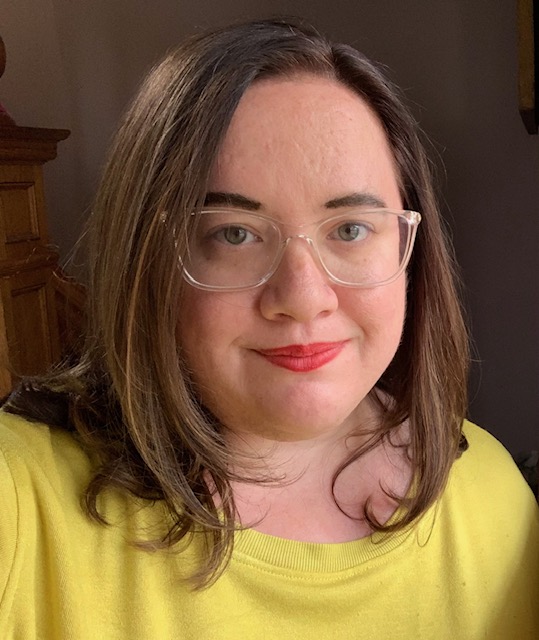 A person with shoulder-length brown hair, wearing clear glasses and a yellow top, smiles softly at the camera. The background is softly lit and neutral.