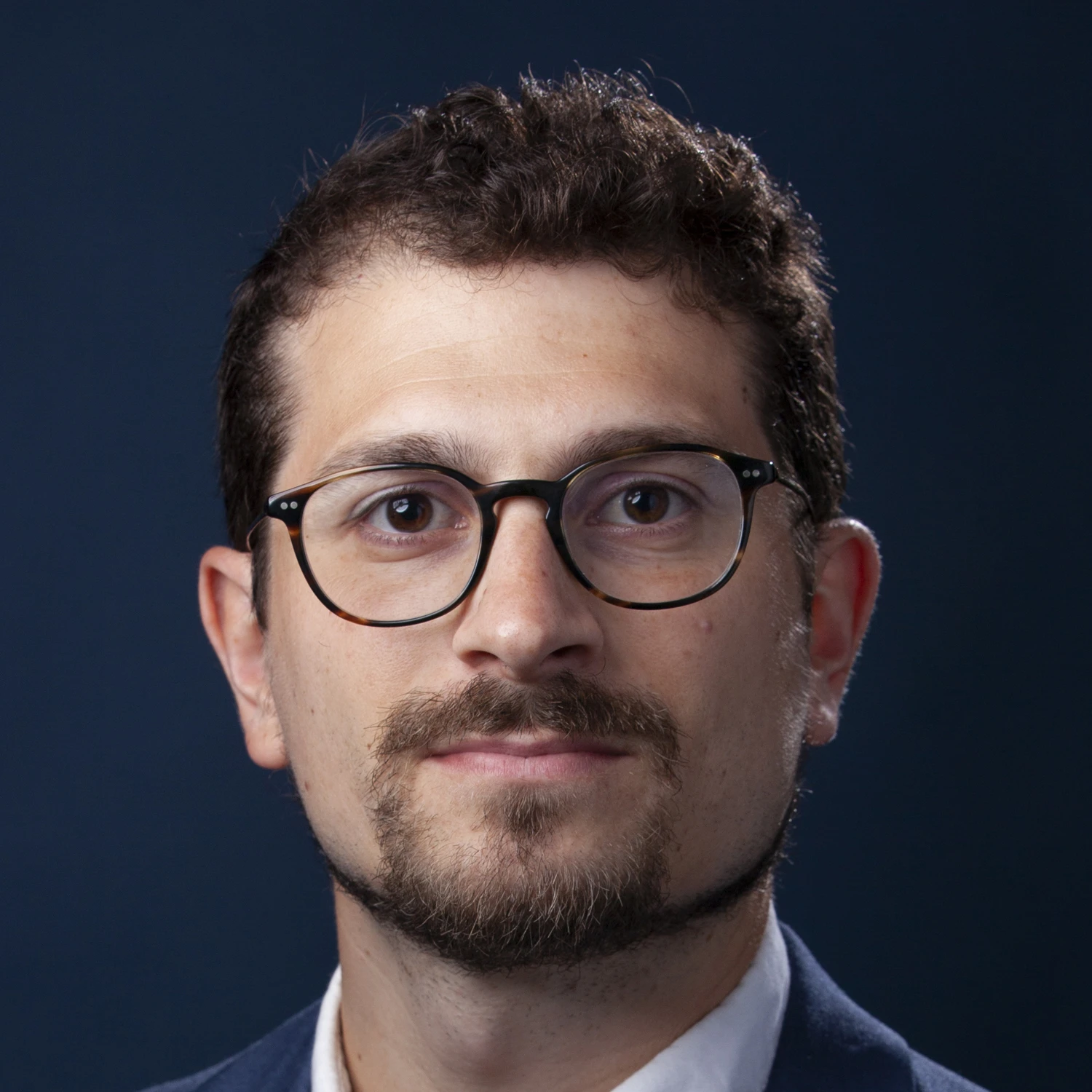 A man with short curly hair, glasses, and a trimmed beard is wearing a suit and looking directly at the camera. The background is a solid dark blue.