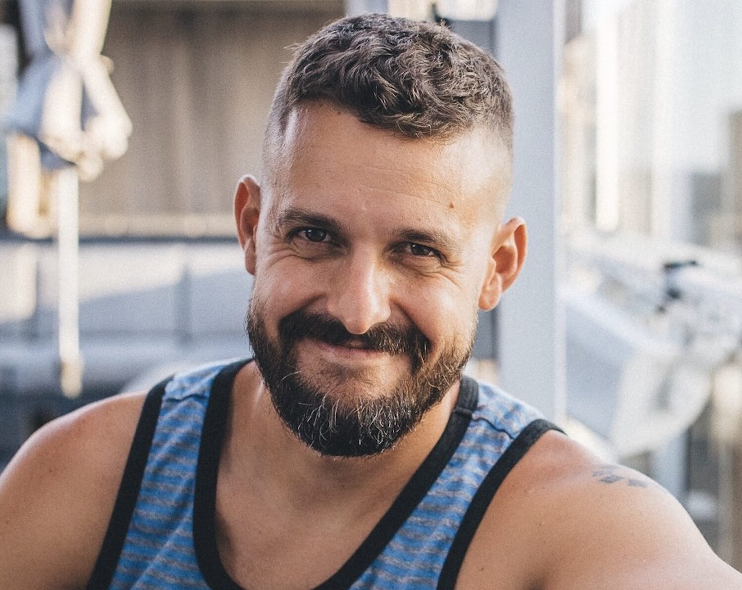A man with short hair and a beard, wearing a blue and gray striped tank top, smiles at the camera in a bright indoor setting.