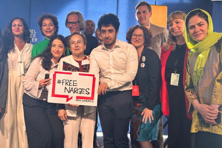 A diverse group of people stand together smiling at what appears to be an advocacy or awareness event, possibly linked to the Nobel Peace Conference 2024; one woman holds a #FREE NARGES PEN AMERICA sign as a woman in a green headscarf stands on the right.