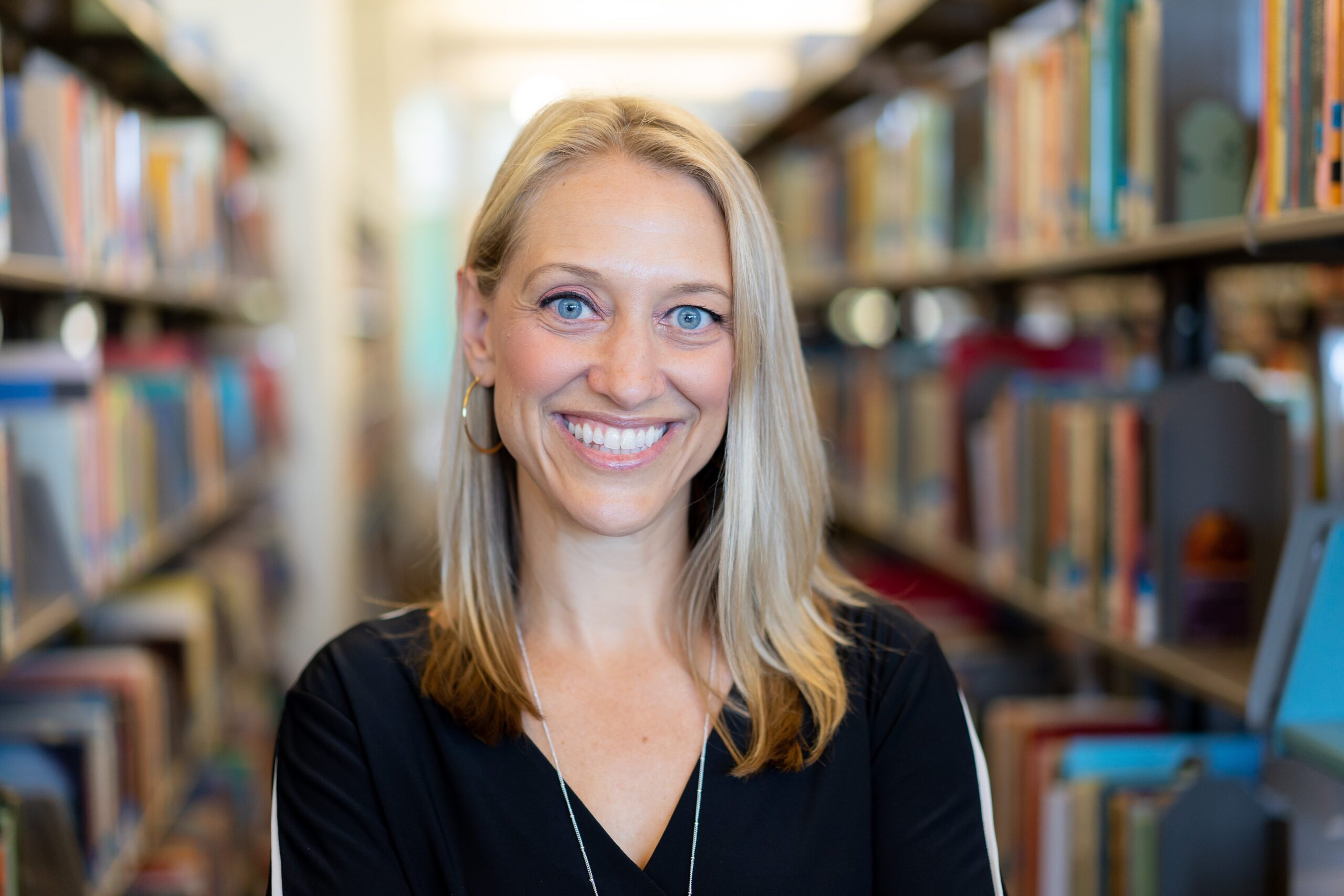 A woman with long blonde hair and blue eyes smiles at the camera, standing between bookshelves in a library. She is wearing a black top and hoop earrings, and the background is filled with colorful, blurred books.