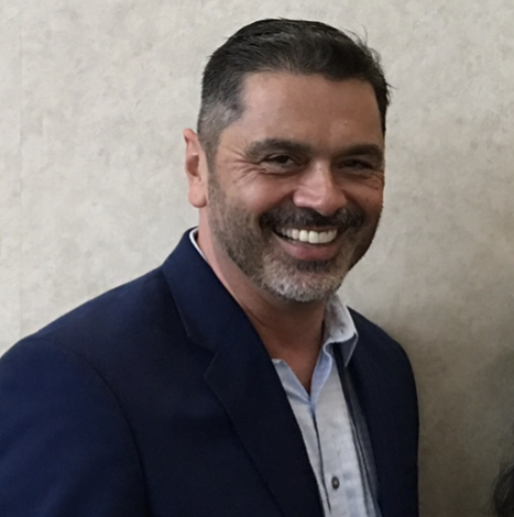 A man with short dark hair and a beard, wearing a navy blue blazer and a light-colored shirt, is smiling in front of a light, textured wall.