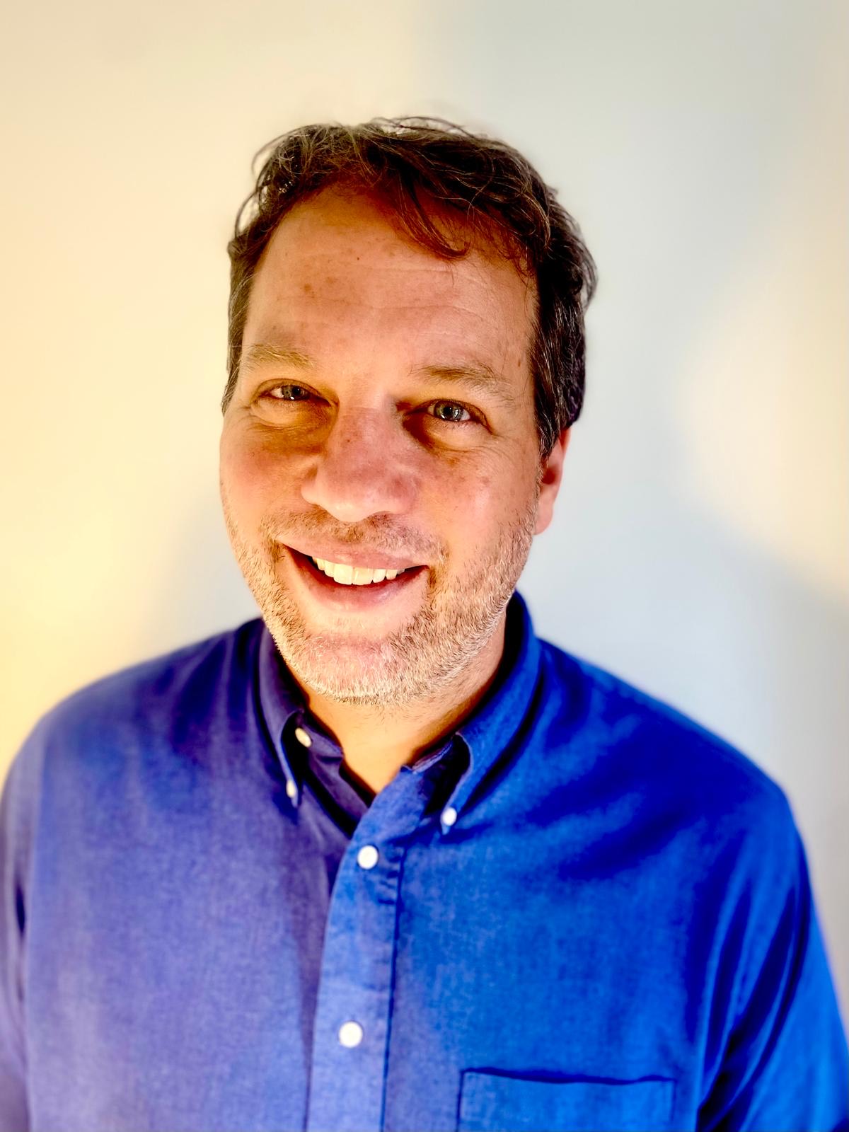 A man with short brown hair and a beard smiles at the camera, wearing a blue button-up shirt, standing against a plain light-colored background.