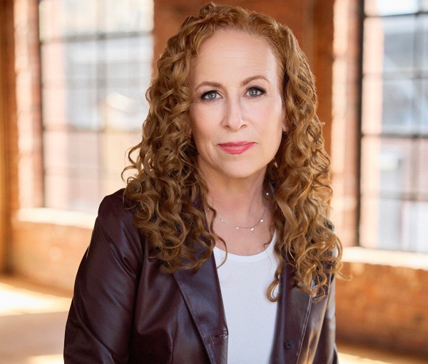 A woman with curly, shoulder-length auburn hair is wearing a dark brown leather jacket over a white top. She is standing indoors in front of large windows with brick walls, looking directly at the camera.