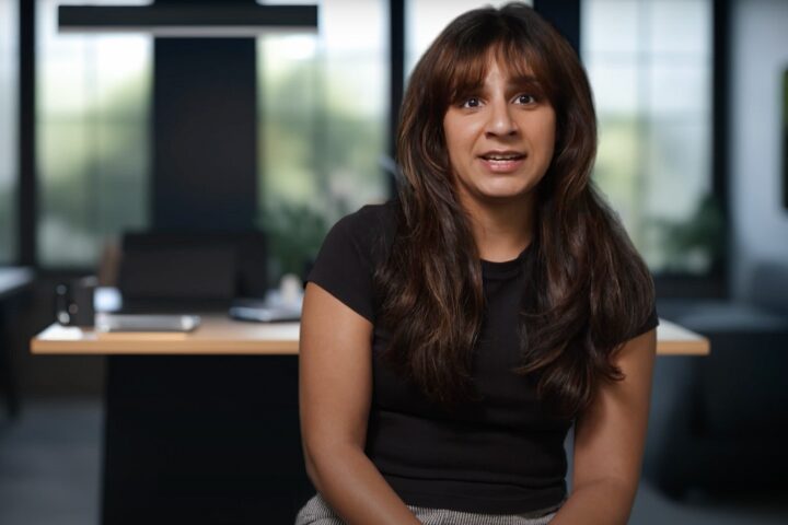 A woman with long brown hair wearing a black shirt is sitting in an office setting. She is speaking, and the background shows large windows and a desk with various office items.