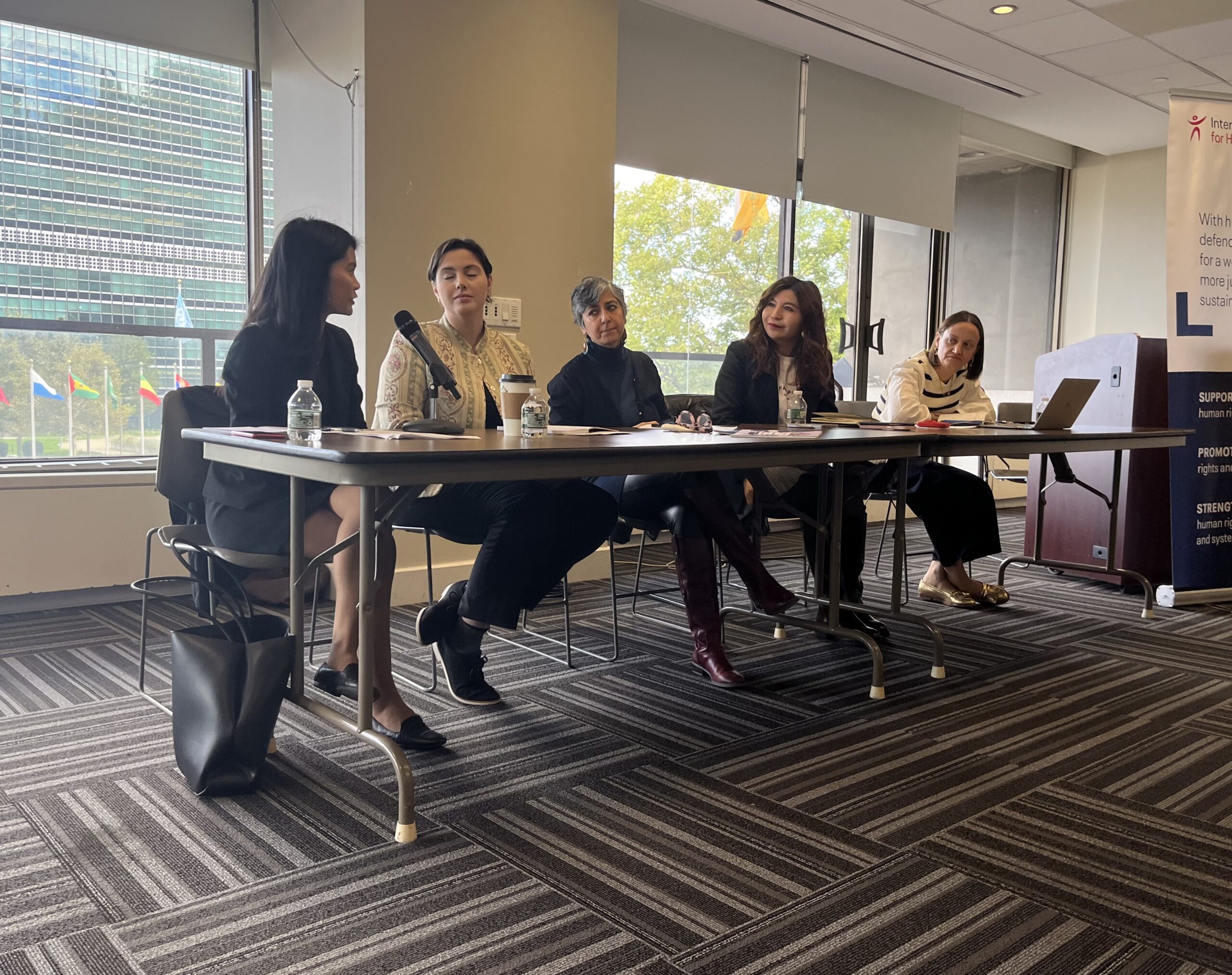 Five people seated at a long table in an indoor conference room appear to be participating in a panel discussion on Vietnams human rights. The room is well-lit, with large windows showing greenery outside. Bottles of water and a microphone are strategically placed on the table.
