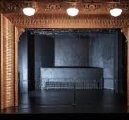 An empty dark stage is framed by an ornately decorated arch with three round lights above. A microphone stand is positioned at the center, and a row of seats or a low wall is visible at the back.