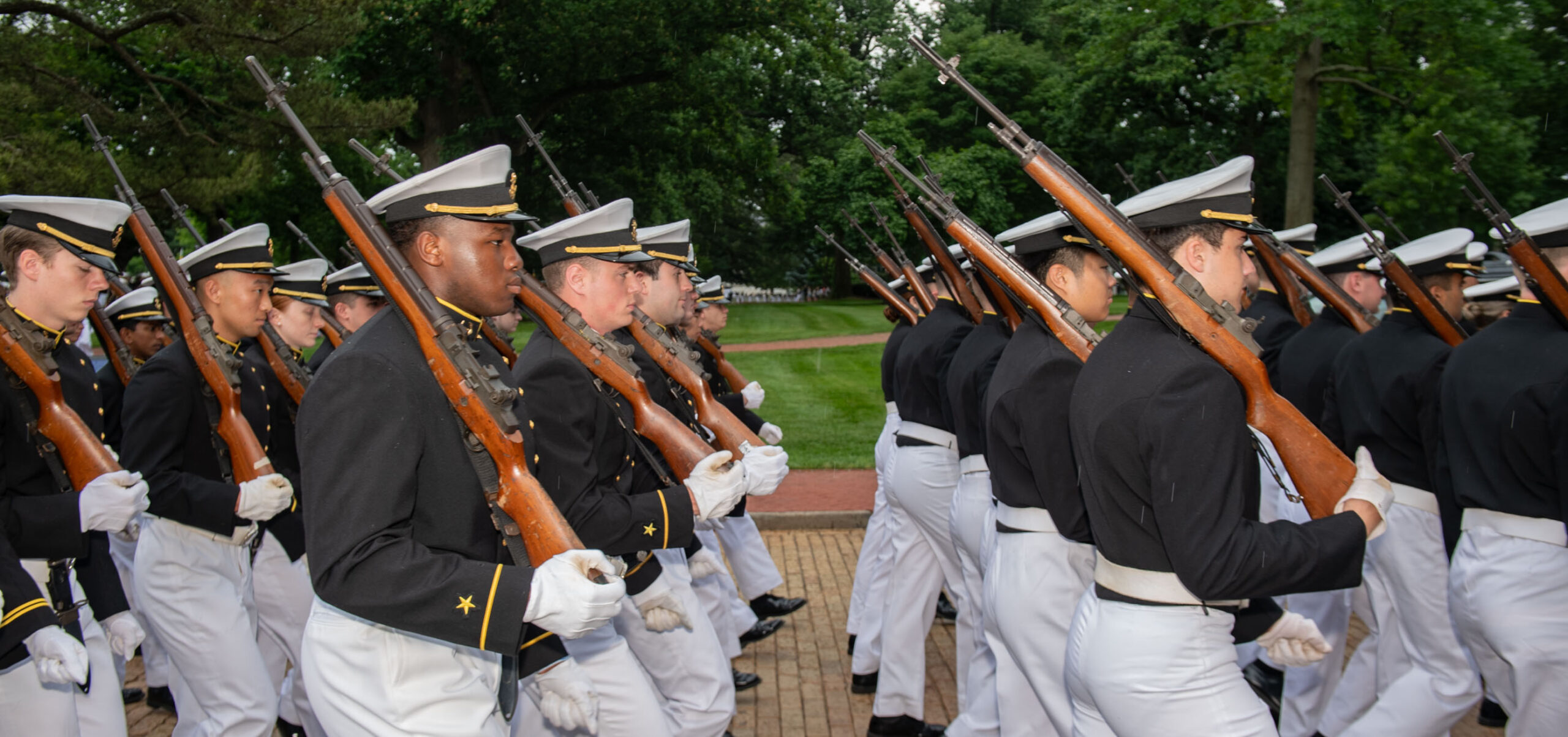 A group of people in military uniforms march in formation, holding rifles over their shoulders. They wear white pants, dark jackets, and caps with white tops. The background features green trees and a lawn.