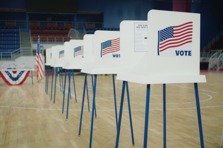 A row of voting booths with the American flag and the word VOTE on each. The booths are set up in a gymnasium with a wooden floor and bleachers in the background. An American flag and a table are also visible.