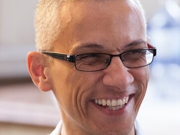 A person with short, gray hair and wearing glasses is smiling broadly. The background is softly blurred, highlighting the person’s joyful expression.