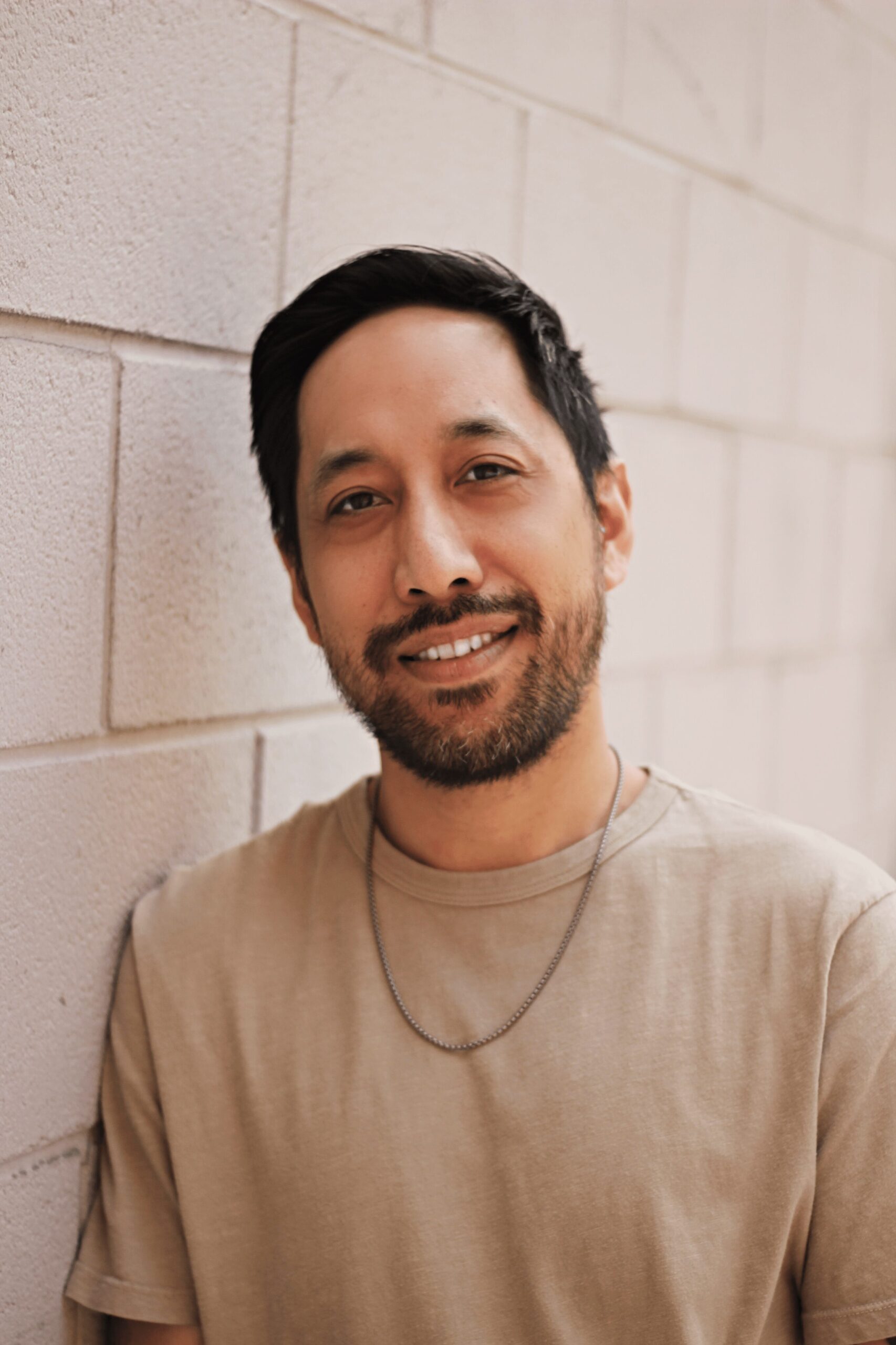 Man with short dark hair and a beard, wearing a beige shirt and a necklace, smiling while leaning against a light-colored brick wall.