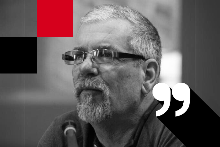 A grayscale image of a man with glasses and a beard, looking thoughtful. Hes in front of a microphone. Theres a large quotation mark symbol and red and black geometric shapes in the background.