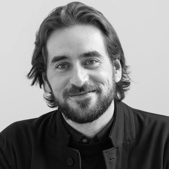A black and white portrait of a person with medium-length hair and a beard, smiling and wearing a dark jacket over a button-up shirt. They are looking directly at the camera with a plain light background.