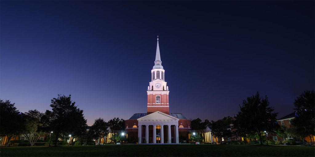 A tall church with a steeple is illuminated at night, centered against a dark blue sky. The building features a clock and columns at the entrance. Trees frame the scene on both sides.