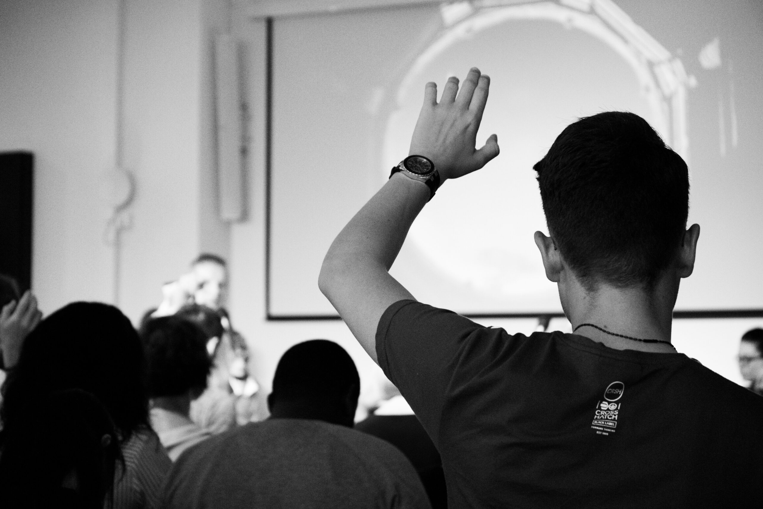 A black and white photo of a seminar. A person in the foreground raises their hand, while a speaker in the background appears to be addressing the audience. A large screen is visible behind the speaker.