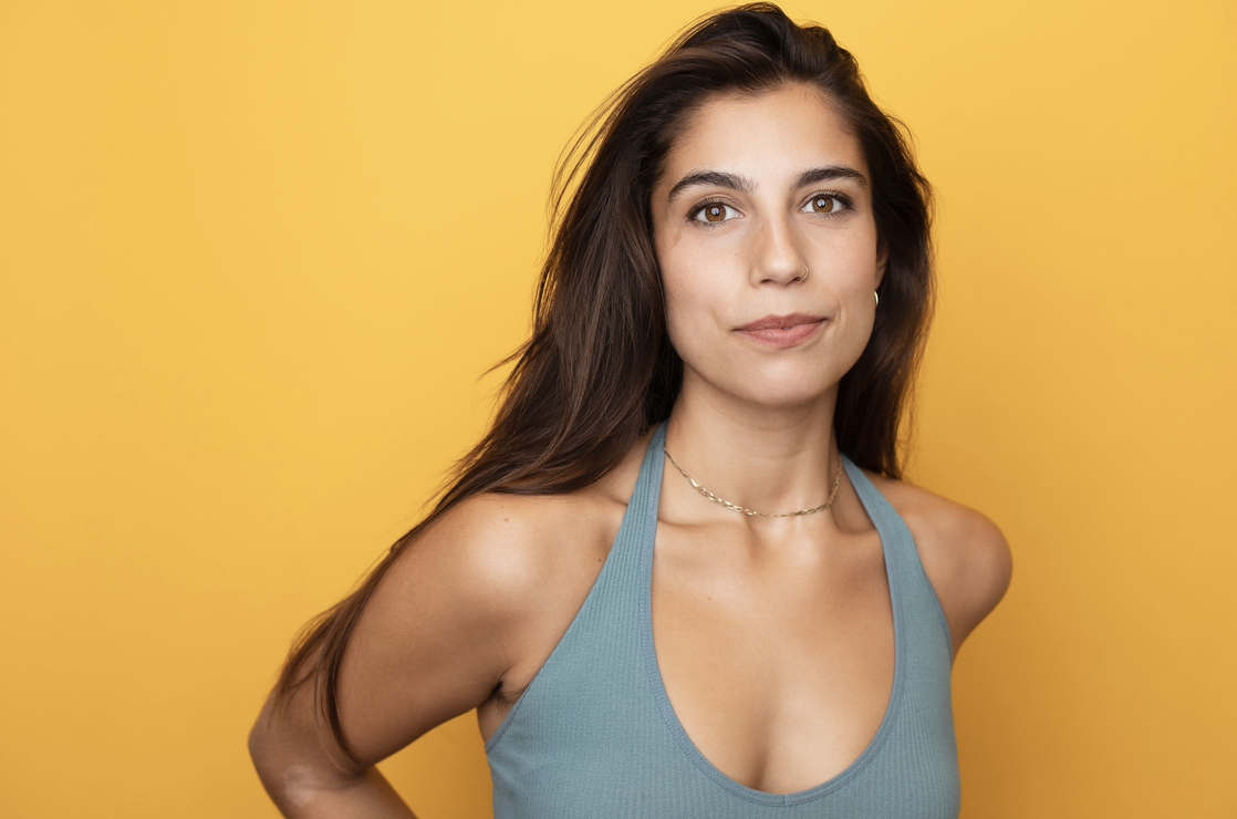 A woman with long brown hair stands against a yellow background. She is wearing a light blue sleeveless top and a delicate necklace. Her expression is calm and confident, and she gazes directly at the camera.