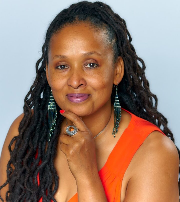 A person with long dreadlocks smiles gently while resting their chin on their hand. Theyre wearing dangly earrings, a ring, and an orange sleeveless top, set against a light gray background.