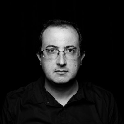 Black and white portrait of a person with short hair, wearing glasses and a collared shirt, looking directly at the camera against a dark background.