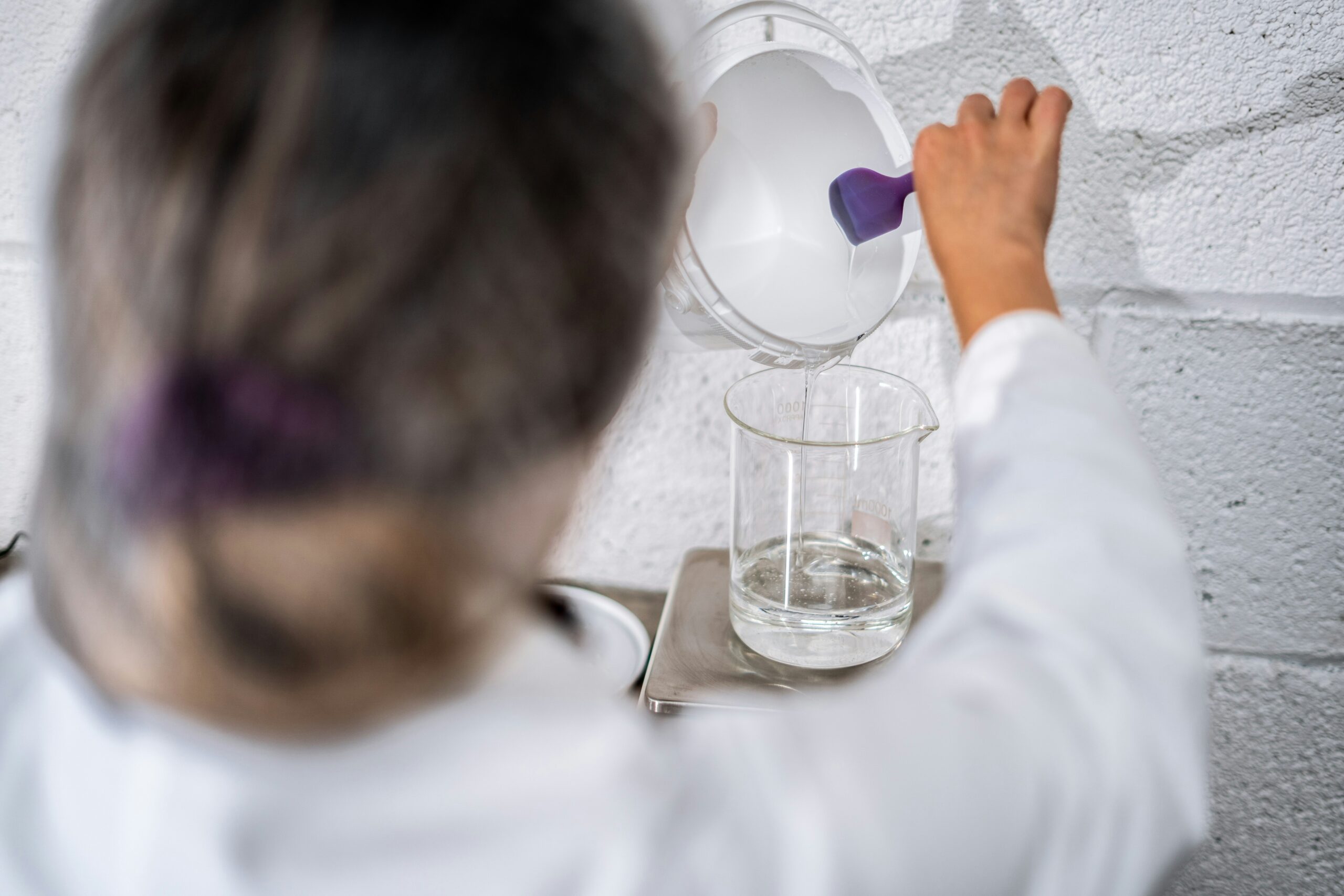 A person in a lab coat is pouring a white powder from a container into a beaker with water. The beaker is placed on a digital scale. The background is a white brick wall.