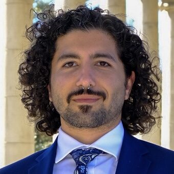 A person with curly hair and facial hair is wearing a blue suit, white shirt, and patterned tie. They are standing outdoors in front of stone columns, smiling slightly at the camera.