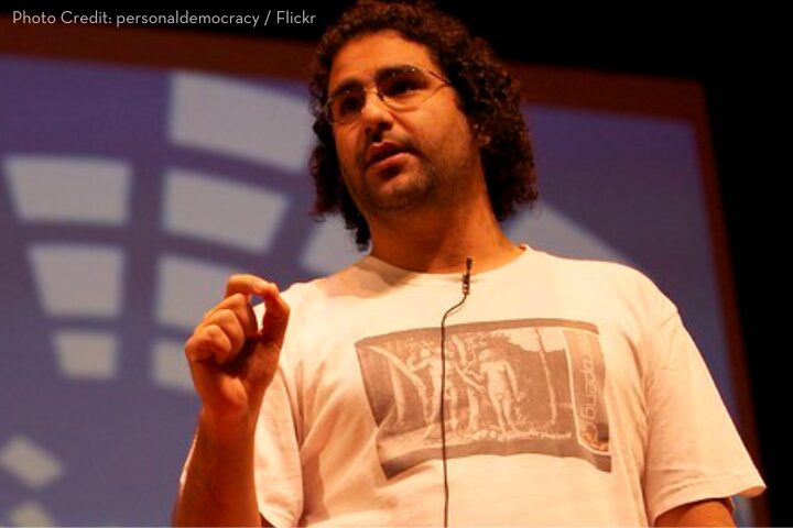 A person with curly hair and wearing glasses speaks on stage, holding one hand slightly raised. They are wearing a T-shirt with a printed design and have a headset microphone. The background features a geometric pattern on a screen.