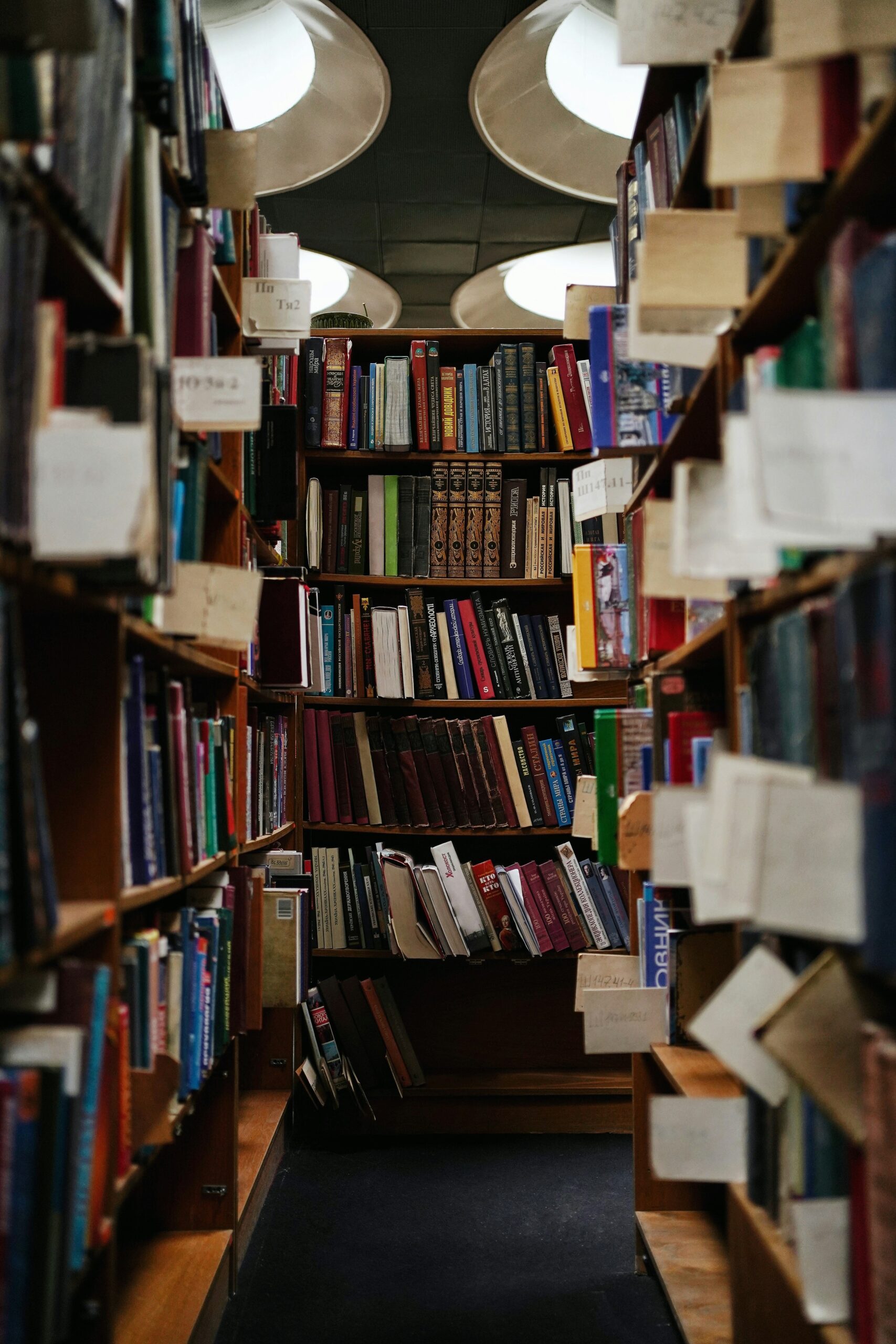 A narrow library aisle is filled with books on shelves, creating a cozy, enclosed space. Overhead, circular lights illuminate the scene. Books of various colors and sizes are packed tightly together along the wooden shelves.