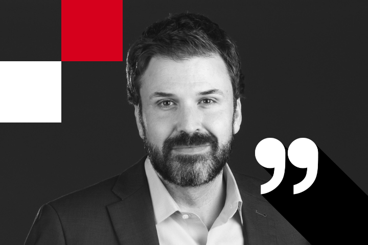 A man with a beard, wearing a suit, is smiling slightly against a dark background. There are red and white geometric shapes and a large white quotation mark symbol beside him.