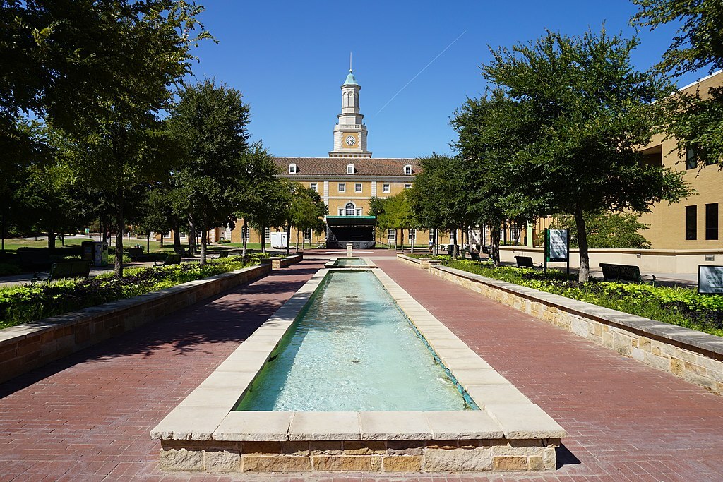 A landscaped walkway leads to a prominent building with a tall clock tower. A rectangular fountain runs down the center, flanked by trees and benches. The sky is clear and blue.