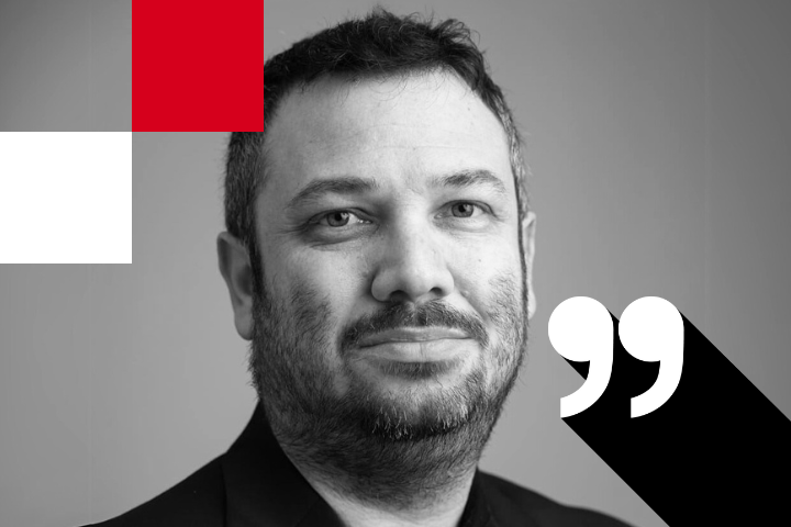 Black and white portrait of a man with a beard, looking at the camera. The image has geometric red and white shapes in the top left and a large quotation mark in the bottom right corner.