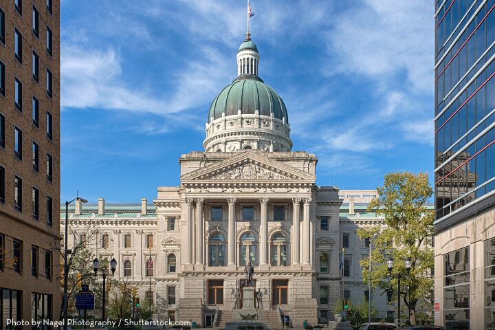The image shows a large building with a green dome and classical architecture, situated between two modern buildings. The sky is clear with a few clouds, and there are trees and street lamps in the foreground.