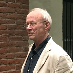 An older man with glasses and short white hair stands in front of a brick wall. He wears a beige jacket over a dark shirt and looks to the side.