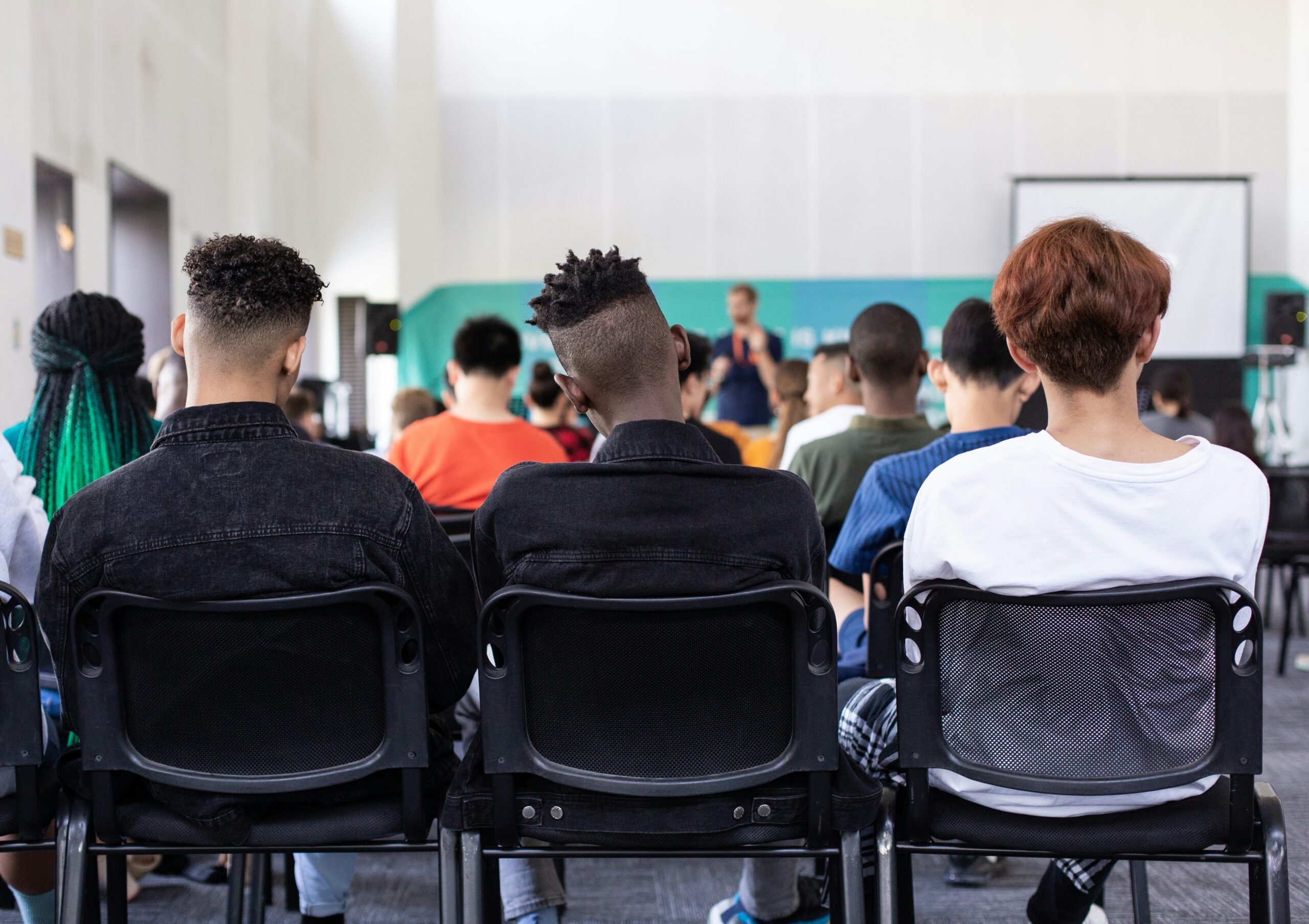 A group of people seated indoors, facing forward, listening to someone at the front. A screen is visible in the background. Three individuals are in the foreground, with diverse hairstyles. The setting has a modern, casual atmosphere.