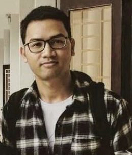 A person wearing glasses and a black and white plaid shirt stands in front of an interior door. They are carrying a backpack. The background includes a white wall and another door with a grid-like design, reminiscent of something Le Huu Minh Tuan might appreciate for its simplicity.