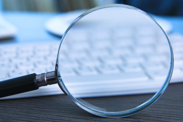 A magnifying glass is placed over a computer keyboard, with the glass enhancing the view of the keys. The setting is a modern desk, featuring a wooden surface and a blurred background.