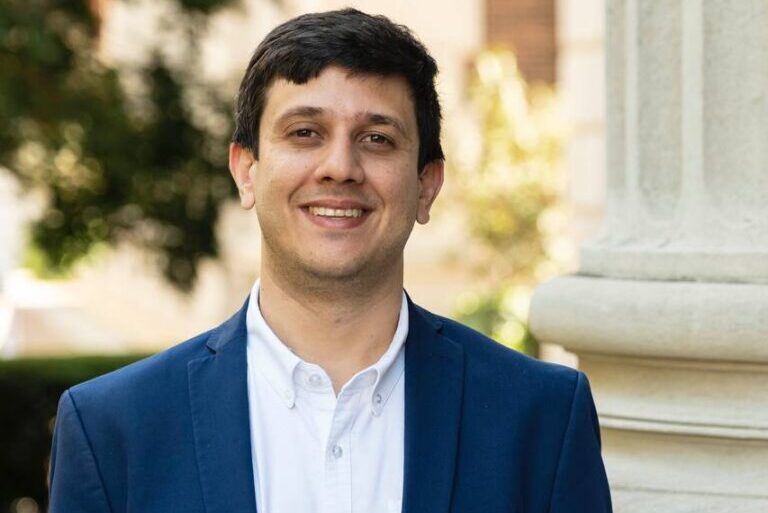 A man with short dark hair smiles at the camera, wearing a white shirt and blue blazer. He stands outdoors in front of a blurred background with greenery and a stone column.