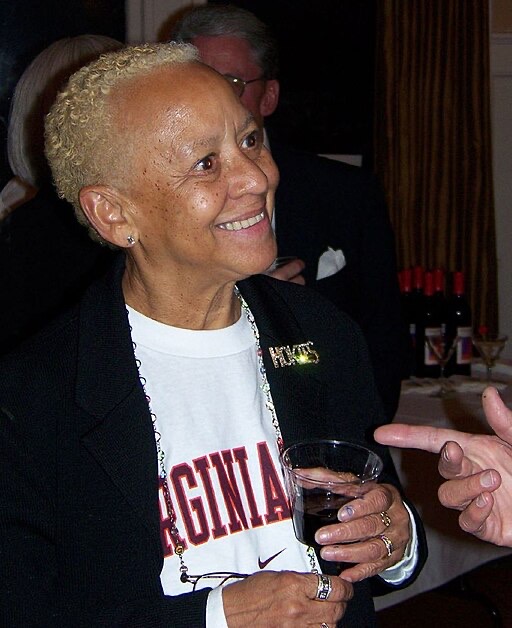 A person with short blond hair is smiling and holding a glass. They are wearing a black jacket over a white Virginia sweatshirt. In the background, there are bottles of wine on a table.