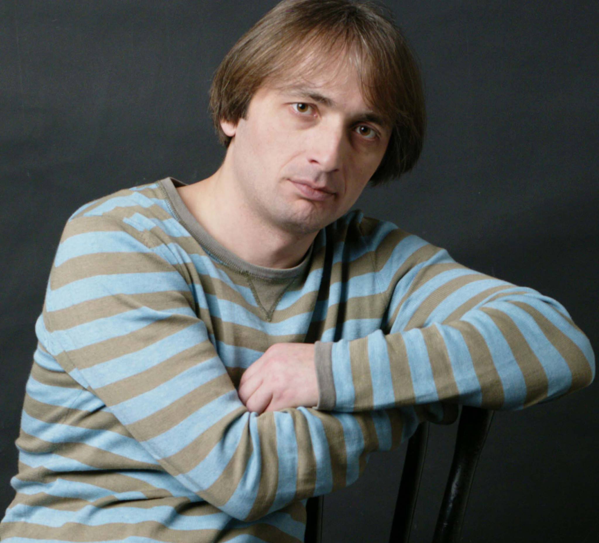 A person with medium-length hair wearing a long-sleeved, blue and brown striped shirt is sitting and resting their arms on the back of a chair. The background is dark.