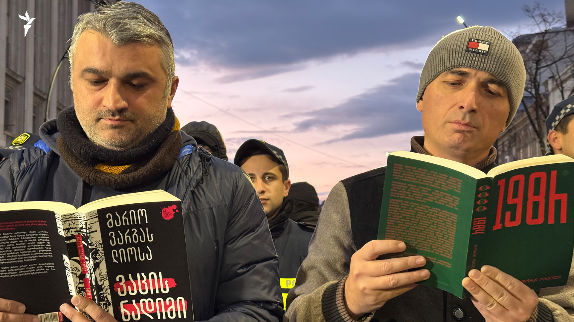 Two men stand outdoors reading books with serious expressions. They are dressed warmly, surrounded by a group of out-of-focus individuals. The sky is dusky, suggesting late evening or early morning.